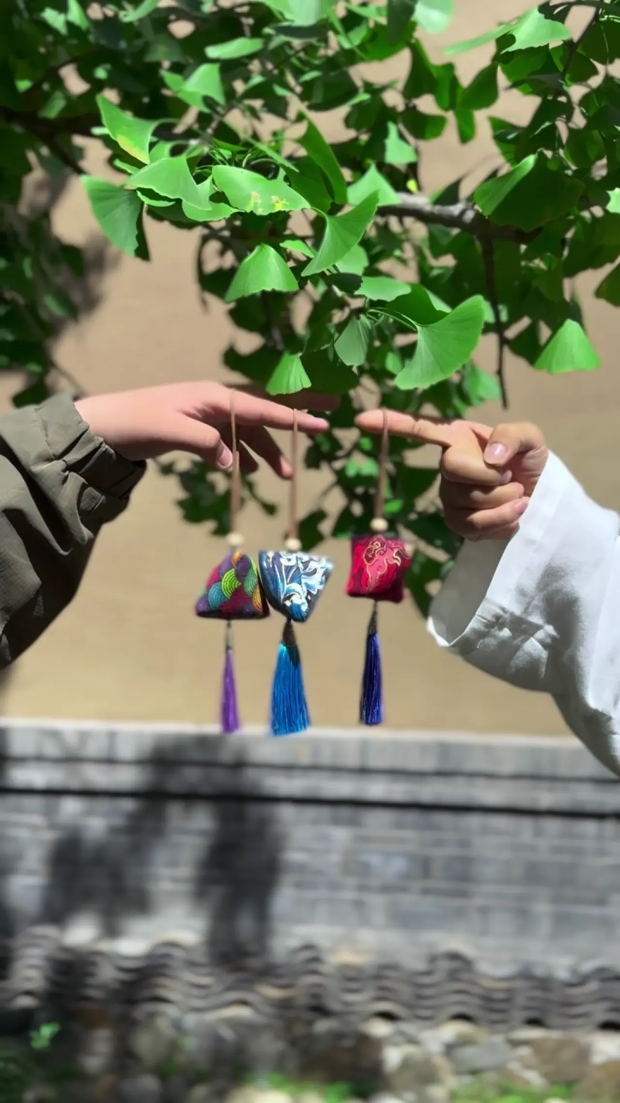 Three embroidered fragrant sachets with purple and blue silk tassels held up beneath a ginkgo branch.