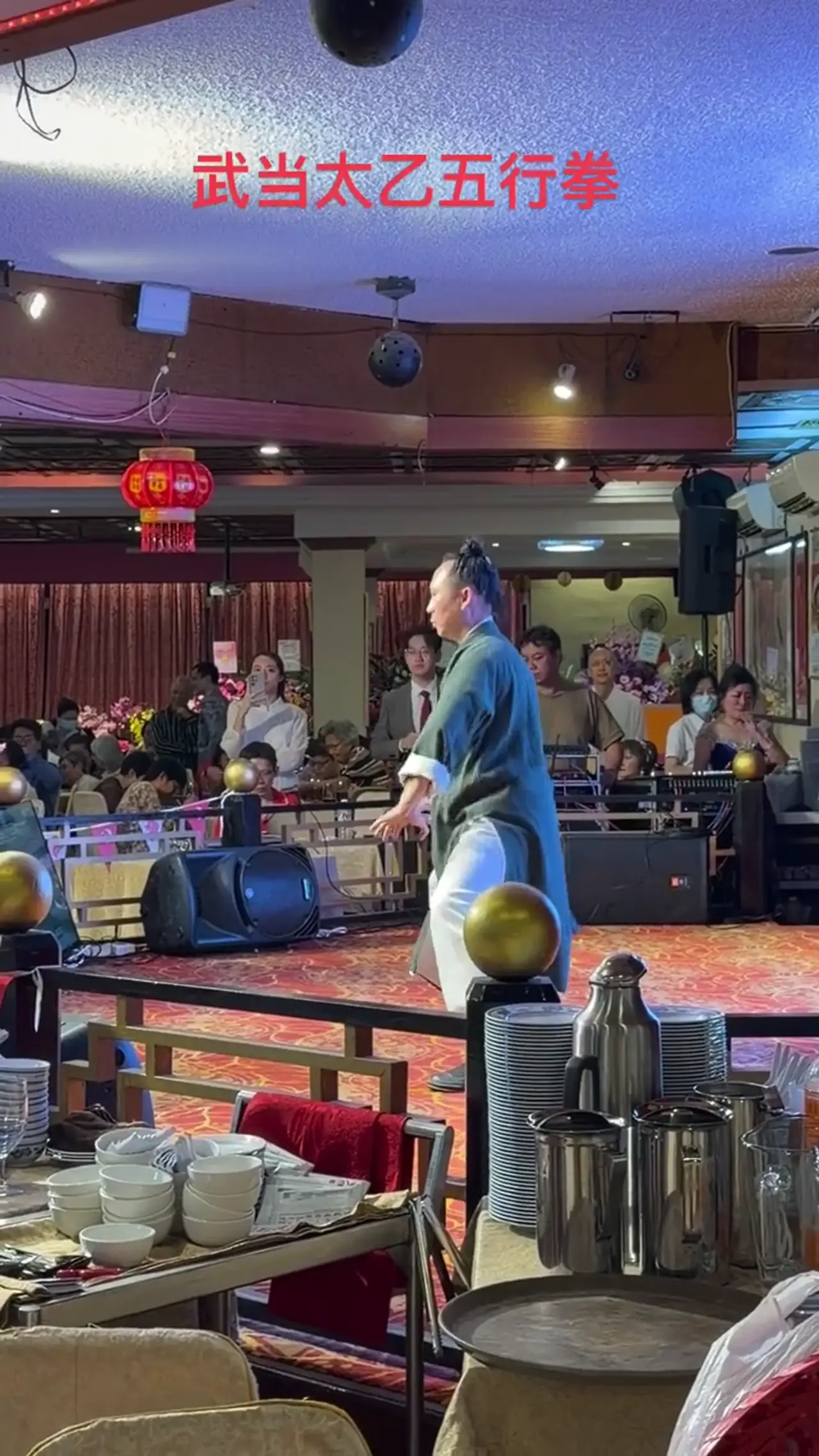 Le maître Yip See Kit, en robe verte, présente le Taiyi des cinq éléments sur une scène de banquet devant les convives.
