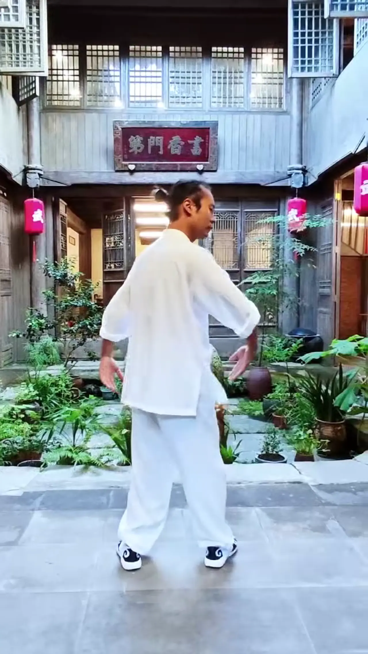 武当-崇恬, vêtu de blanc, pratique le tai-chi dans une cour de bois traditionnel ornée de lanternes rouges et de plantes.