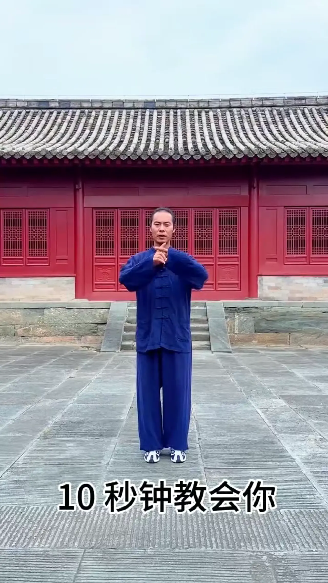 武当-崇恬, en tenue bleue, montre un mouvement dans une cour de pierre devant une salle rouge au toit gris.