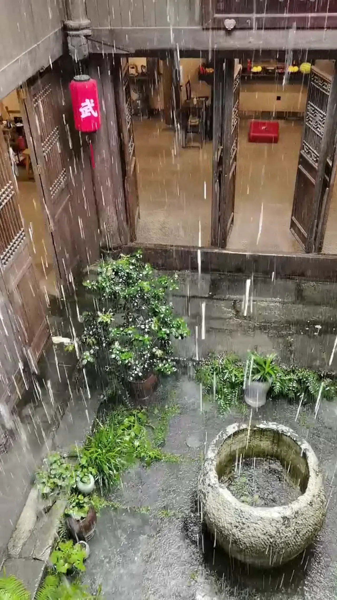 La pluie tombe dans une cour de pierre entourée de bois, avec une lanterne rouge, des plantes en pot et un bassin rond.