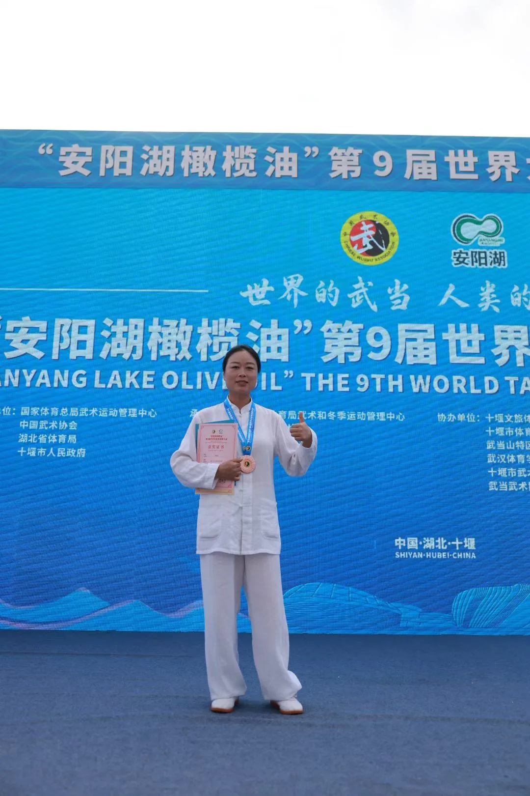 A woman in white wearing a bronze medal gives a thumbs-up.