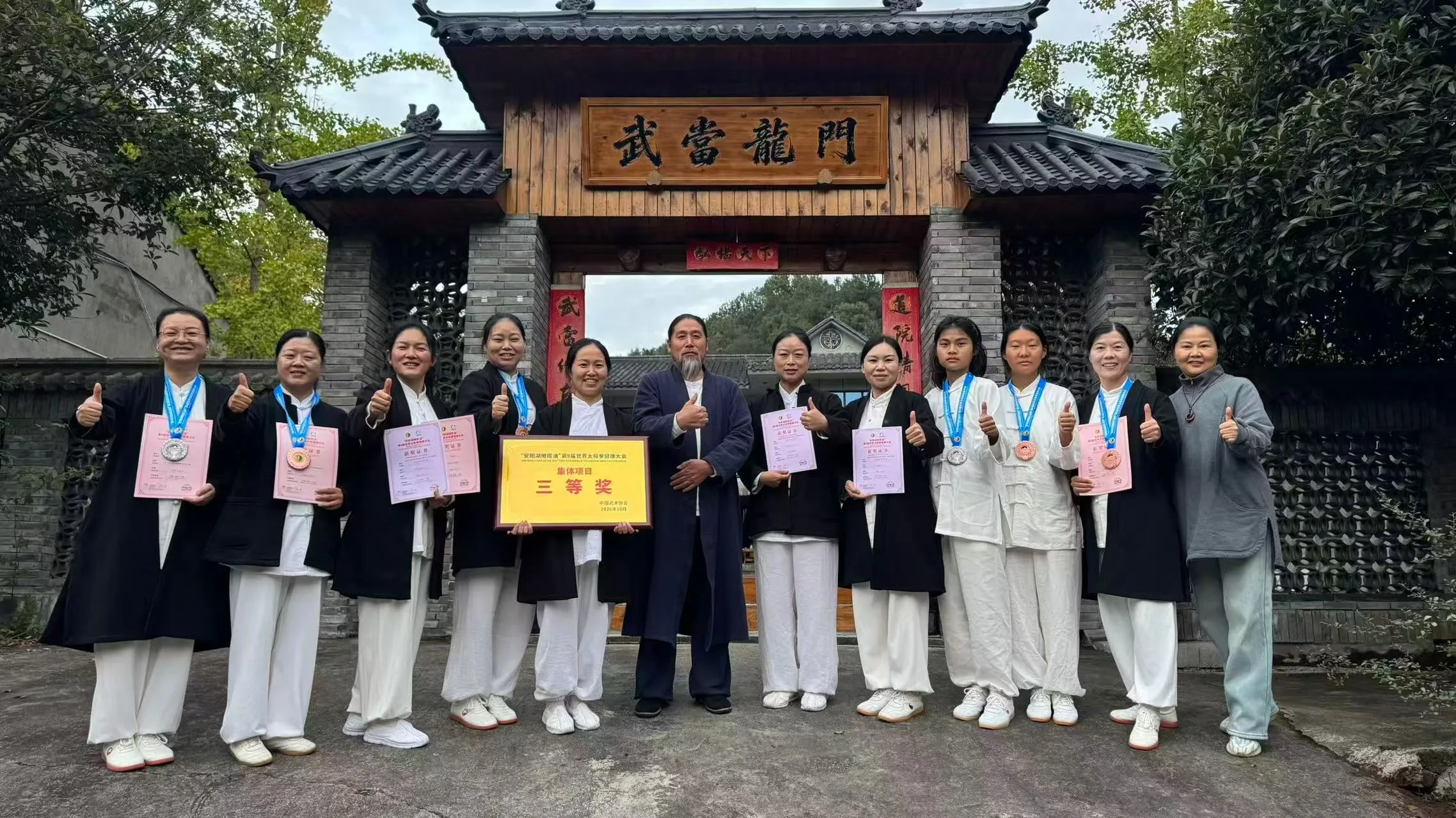 The team in white and black robes holds certificates and a gold plaque before a carved wooden gate reading 武当龍門.