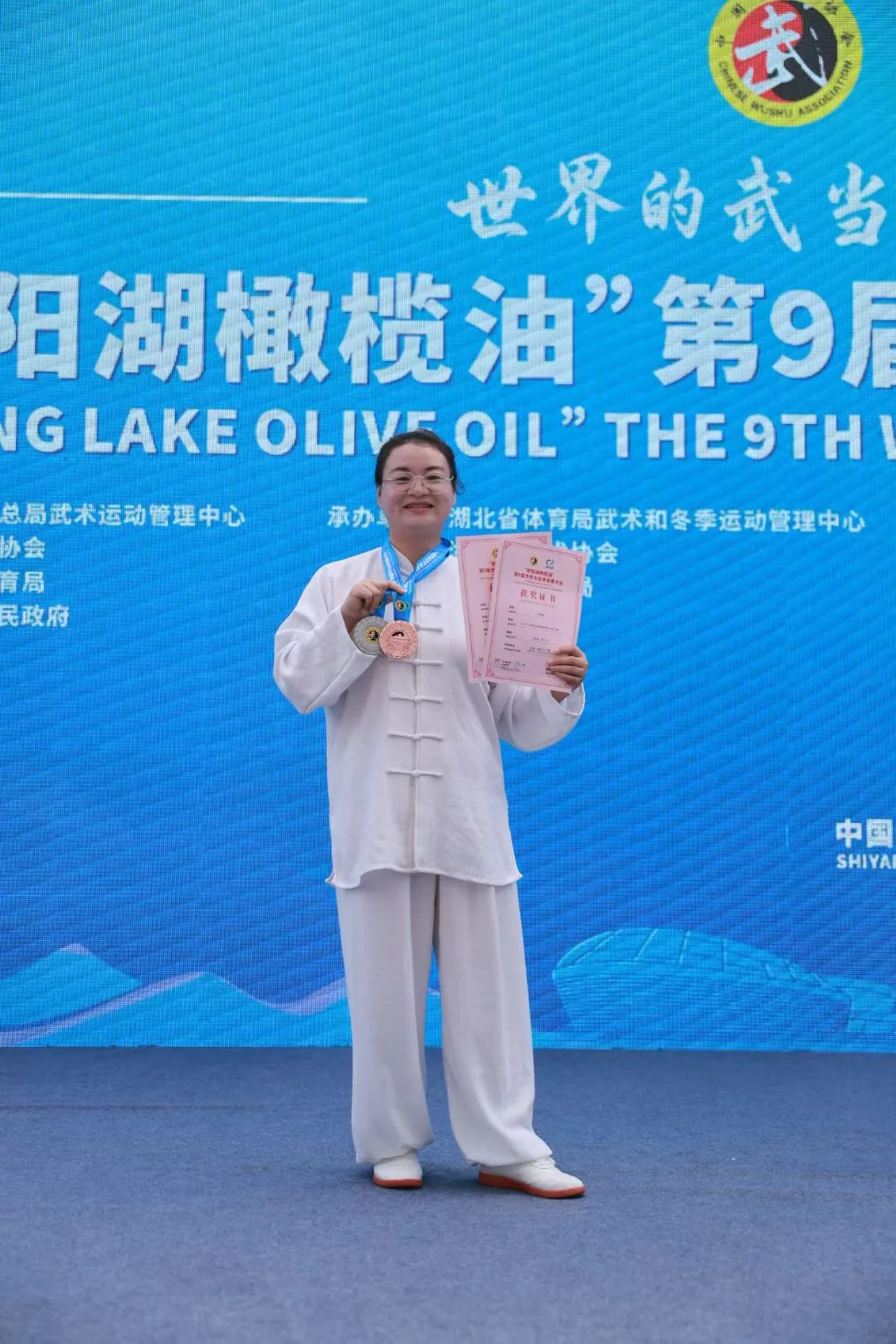 A smiling woman in glasses holds two medals and two certificates before the blue banner.