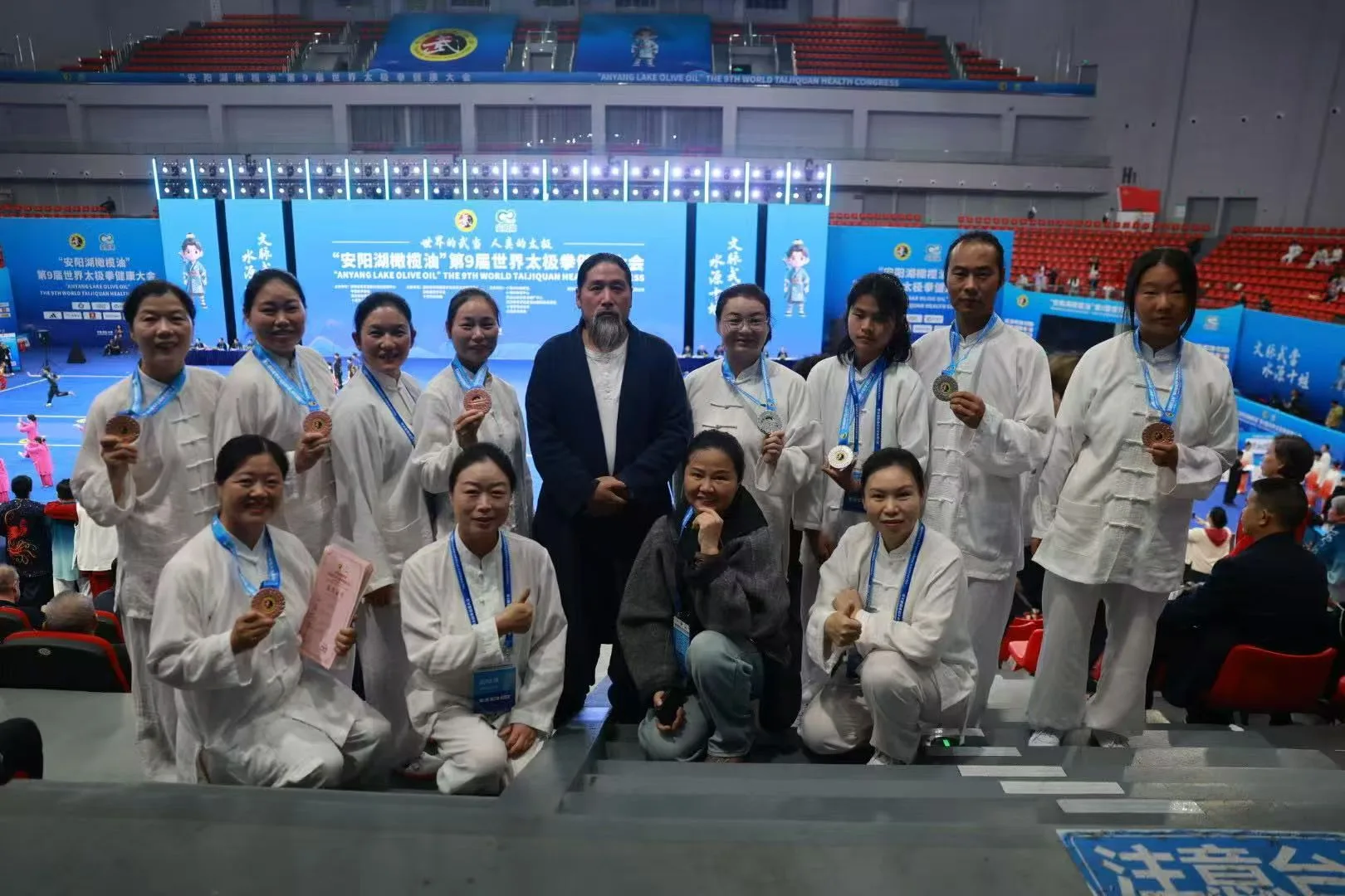 A group in white wearing medals gathers around a bearded teacher in a dark robe, with the arena behind.