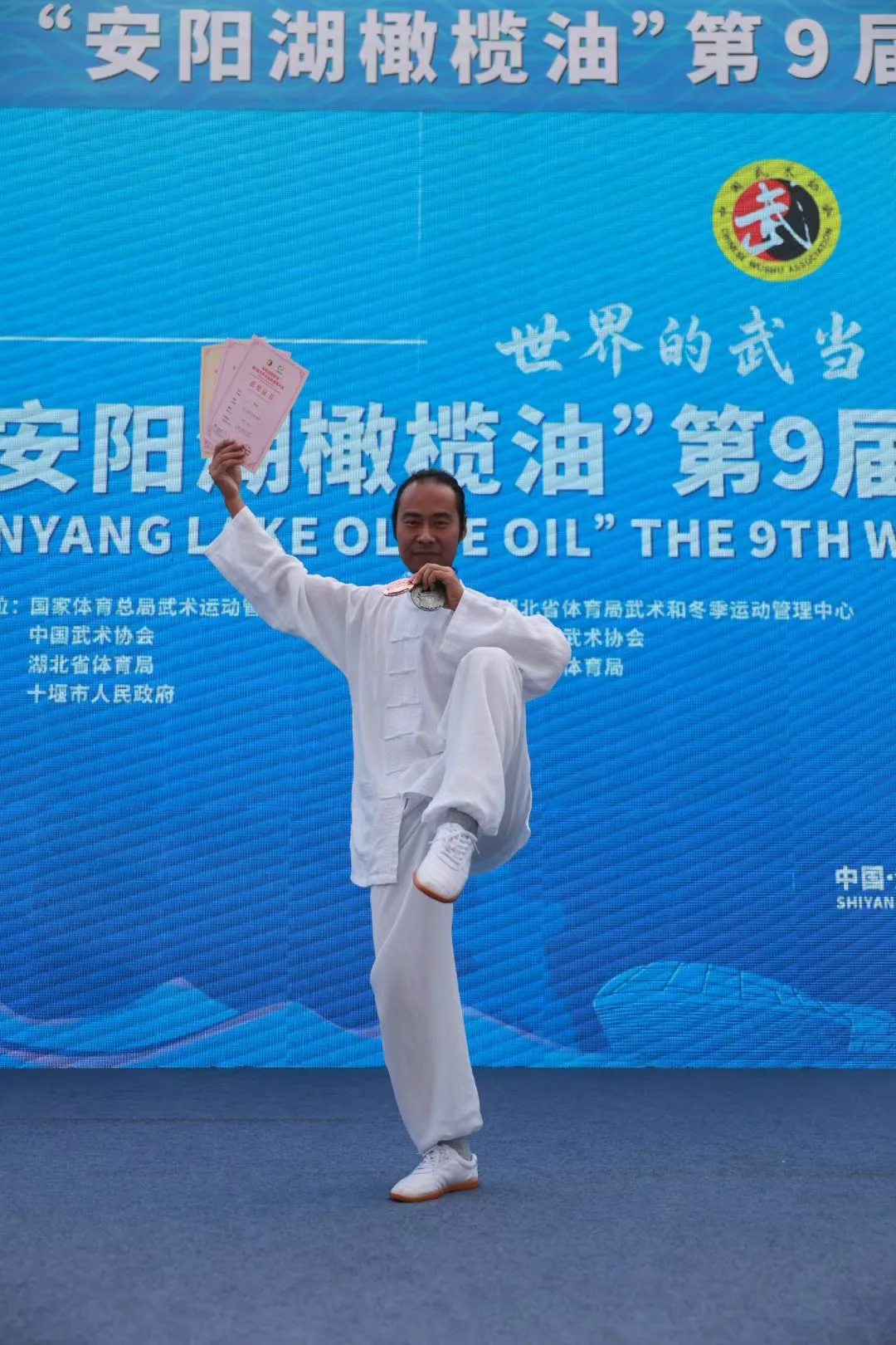 A practitioner balances on one leg, raising award certificates in front of the event banner.
