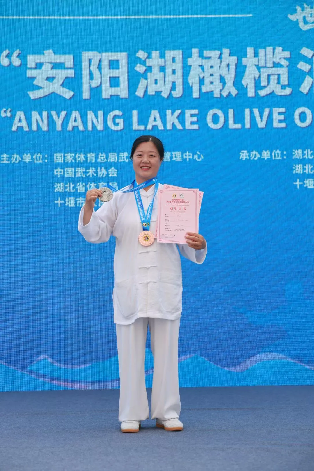 A woman in white holds up a silver medal and a pink award certificate.