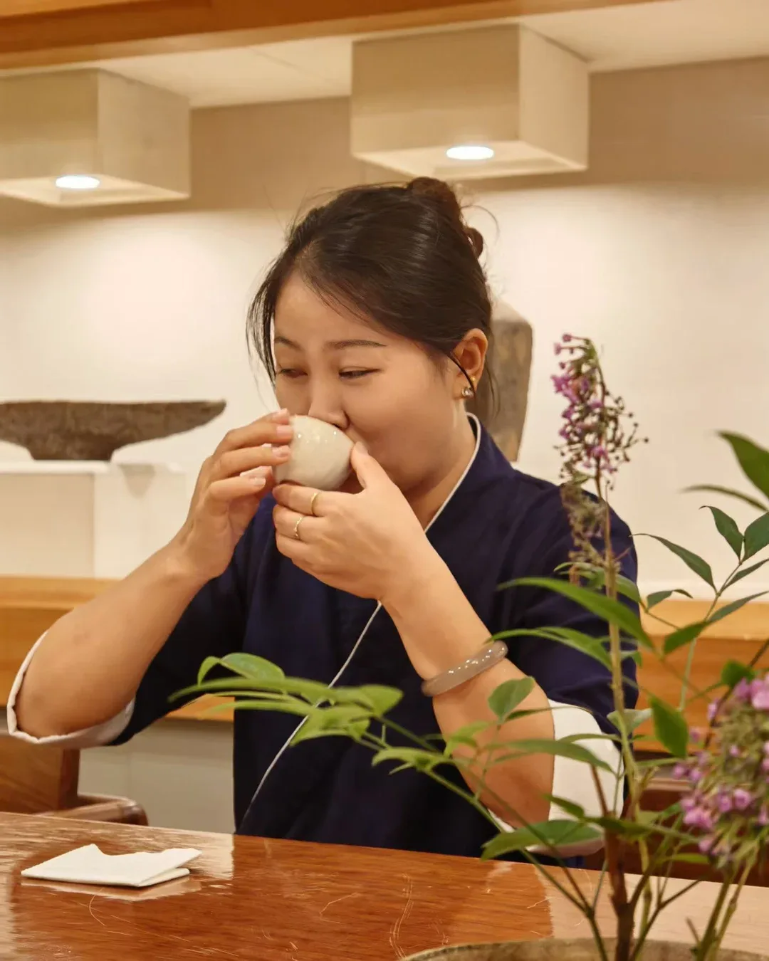 A woman in a navy tea robe holds a small white cup to her lips with both hands, glancing to one side, purple flowers in the foreground.
