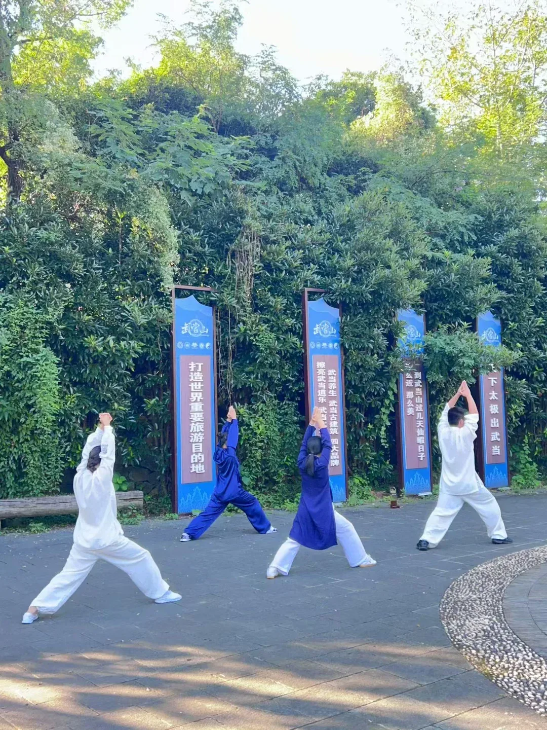 Quatre personnes en blanc et en bleu pratiquent une fente, les paumes jointes au-dessus de la tête, devant des bannières bleues et des arbres.
