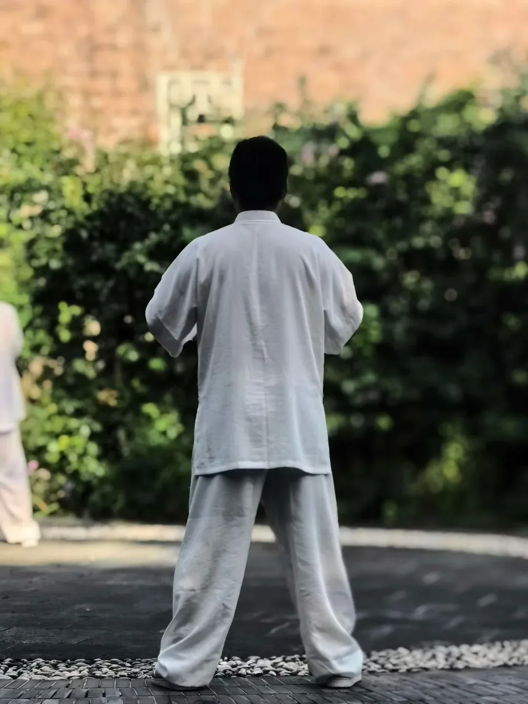 Vu de dos, un homme en tenue blanche se tient les pieds écartés et les bras légèrement avancés, face à une haie verte.