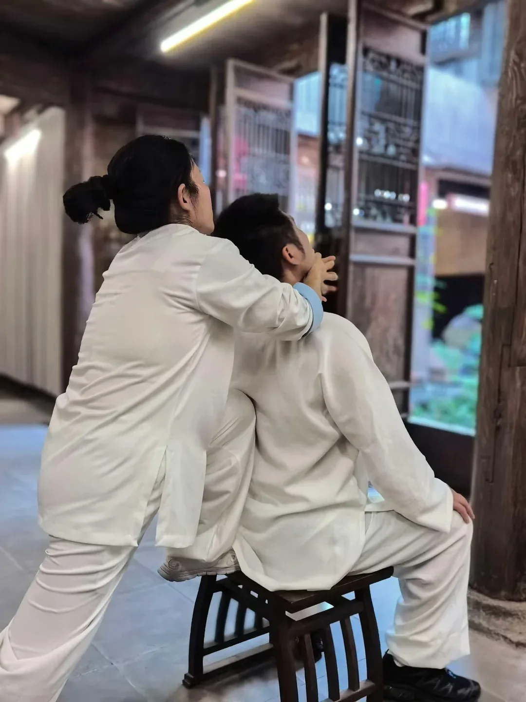 Près de portes à treillis, une femme en blanc accompagne l’étirement d’un homme assis sur un tabouret bas.