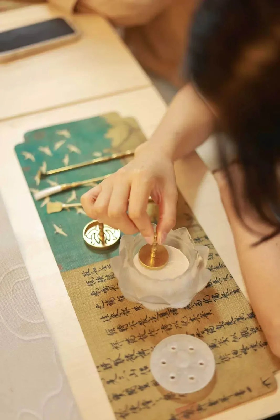 Vue du dessus, une femme abaisse une presse ronde en laiton sur la cendre blanche d’un brûle-parfum en forme de lotus ; les outils reposent sur un tissu imprimé de calligraphie.