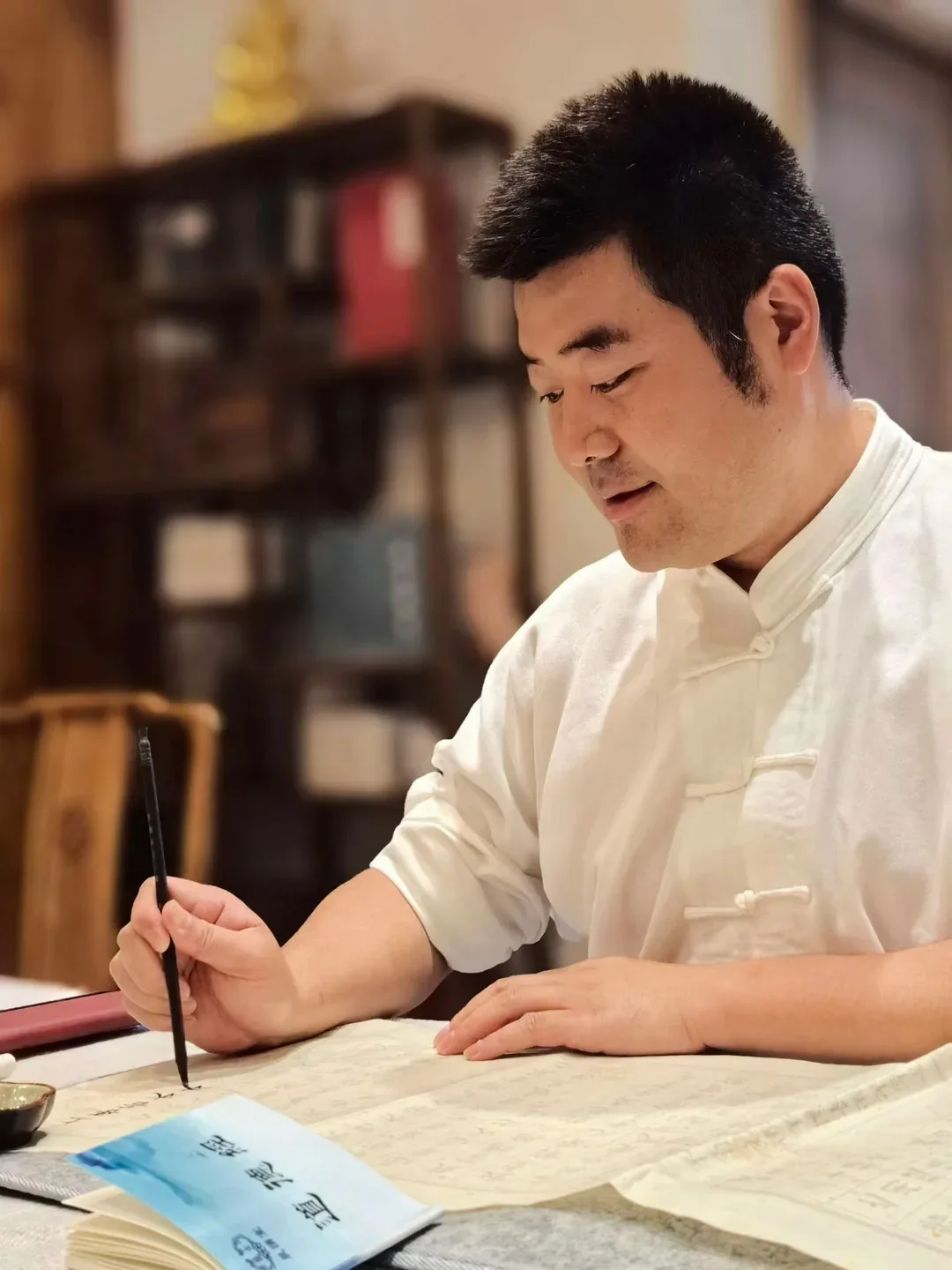 Un homme en veste blanche à boutons noués copie un texte au pinceau, l’autre main sur le papier ; une bibliothèque apparaît derrière lui.