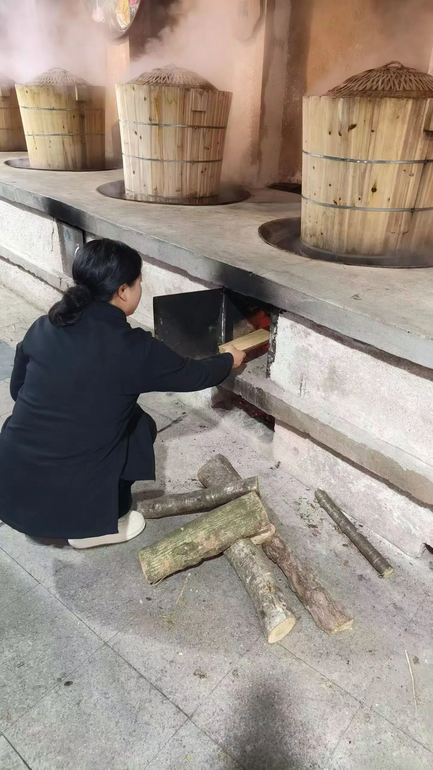 Une personne en robe noire alimente le foyer sous une rangée de cuves en bois d’où s’élève de la vapeur.