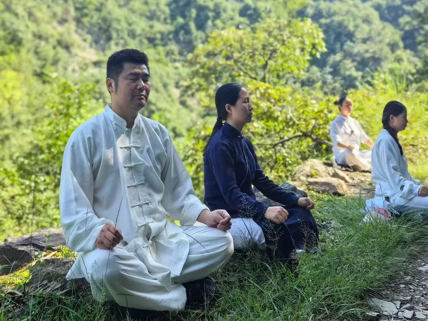 Quatre personnes en blanc et en bleu sombre sont assises, les yeux fermés, sur l’herbe et les rochers près d’un sentier, devant une crête verte et un ciel clair.