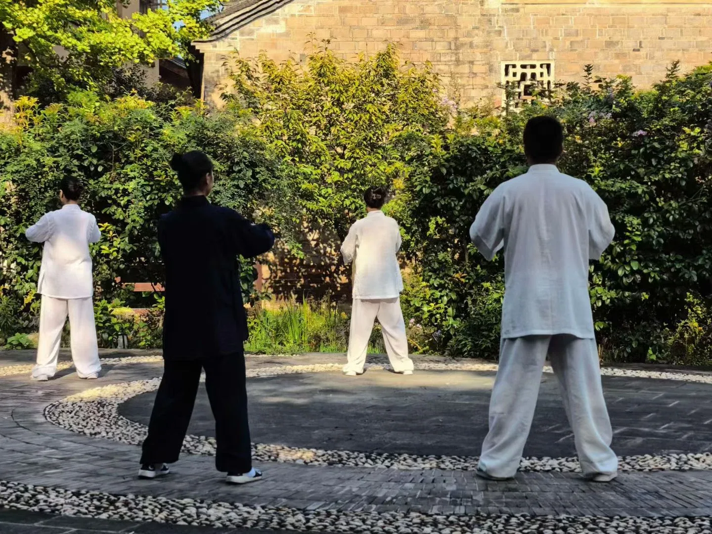 Quatre personnes, trois en blanc et une en bleu sombre, pratiquent dans une cour entourée de galets, face à des arbustes et à un bâtiment ancien.
