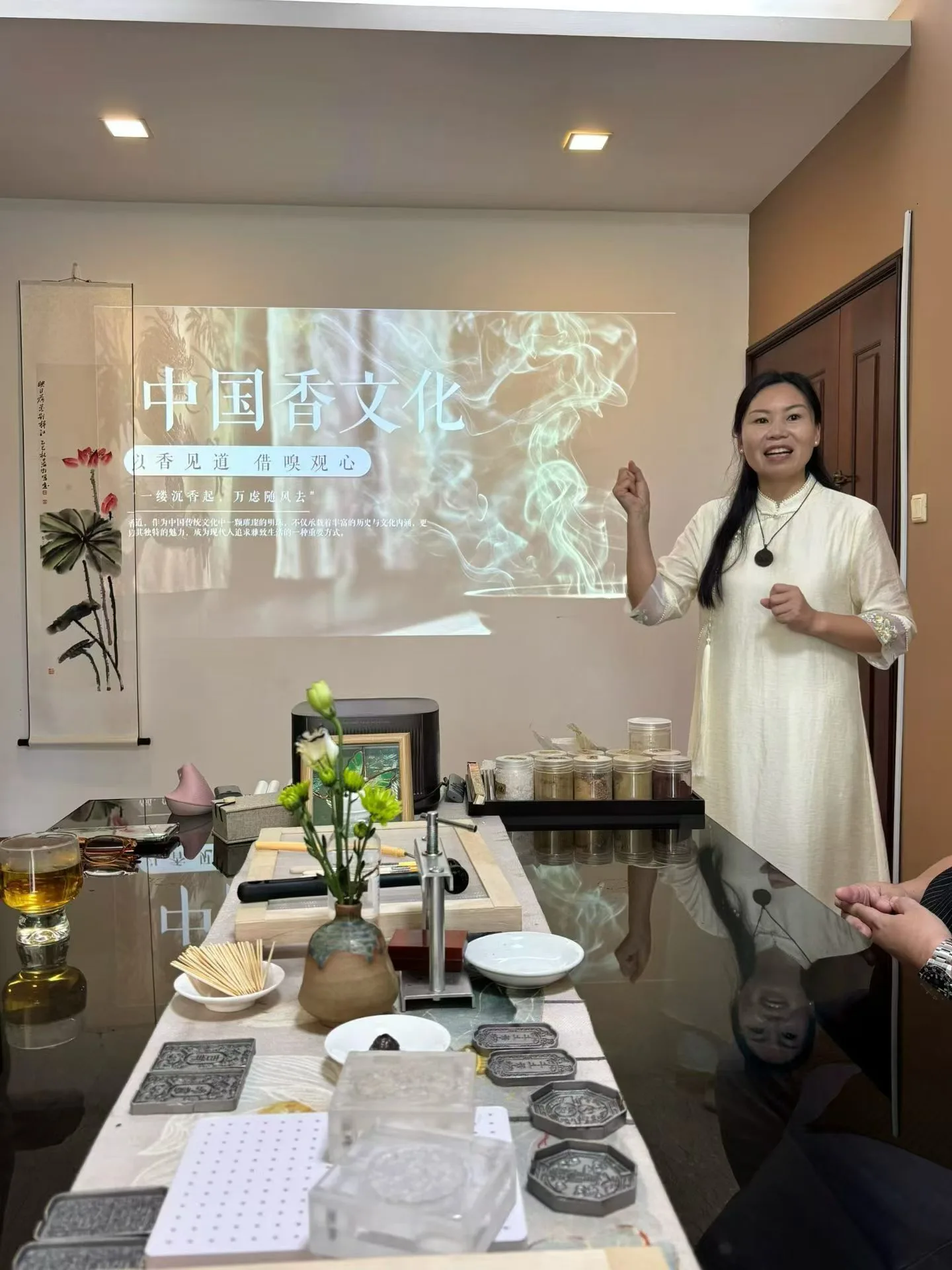 Une enseignante en robe crème parle devant une diapositive intitulée 中国香文化, avec des outils de fabrication d’encens sur la table.