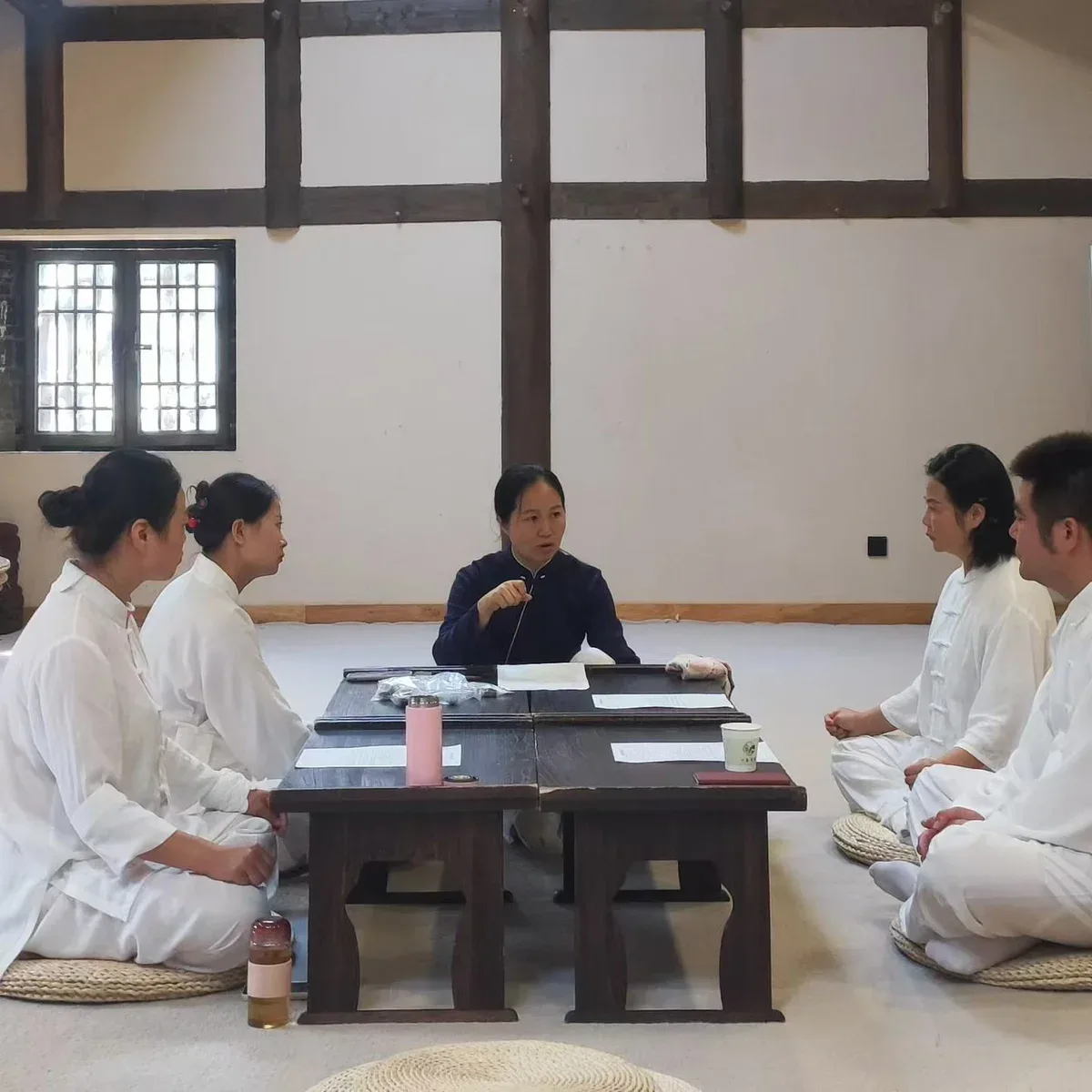 Une femme en bleu sombre parle près d’une table basse, tandis que les participants en blanc l’écoutent dans une salle à poutres apparentes.