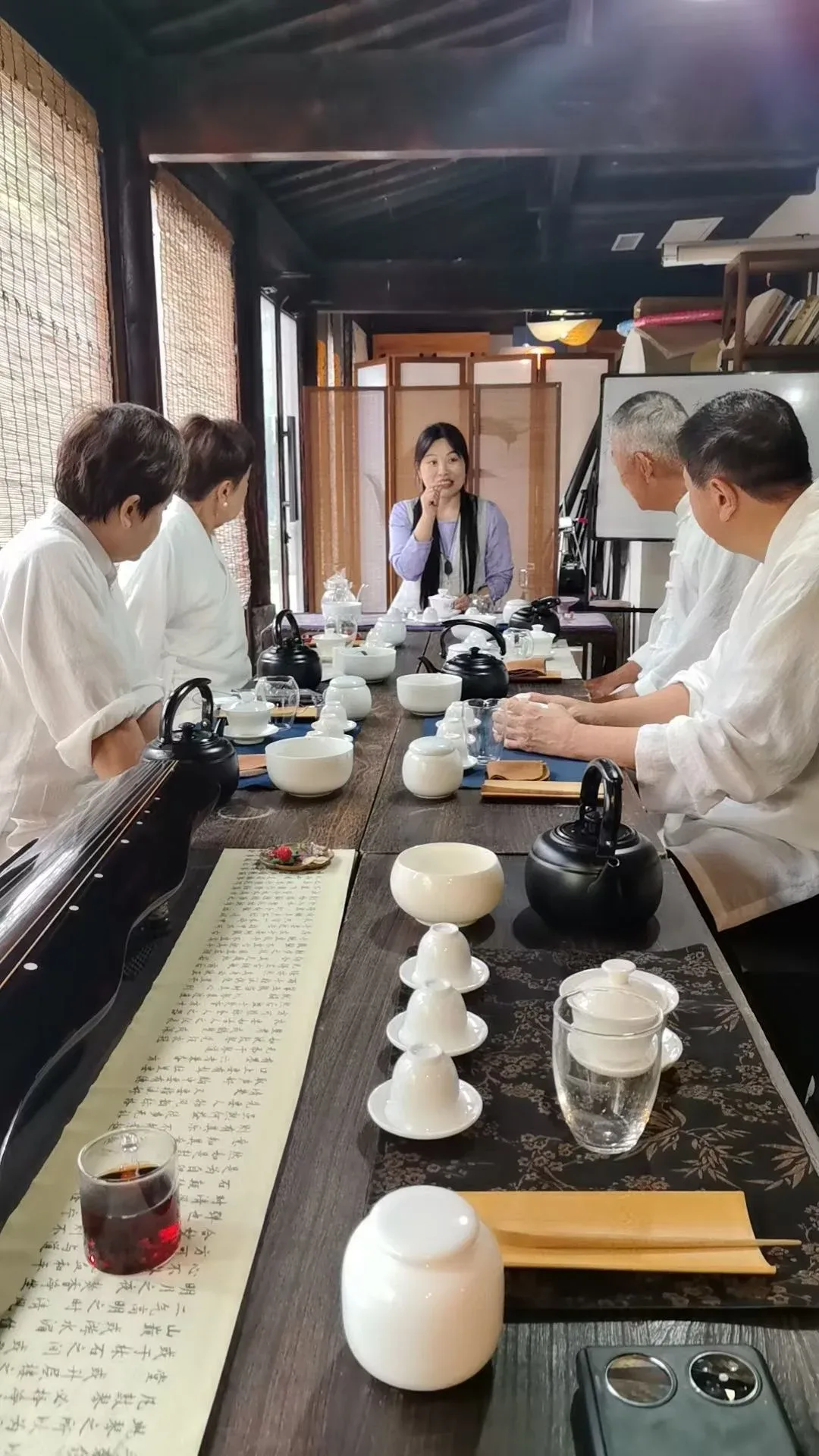 Seen down a long dark table set with white gaiwans, cups and black kettles, a teacher in lilac explains to students seated on both sides; a calligraphy scroll and a glass pitcher of red-brown tea lie in the foreground.