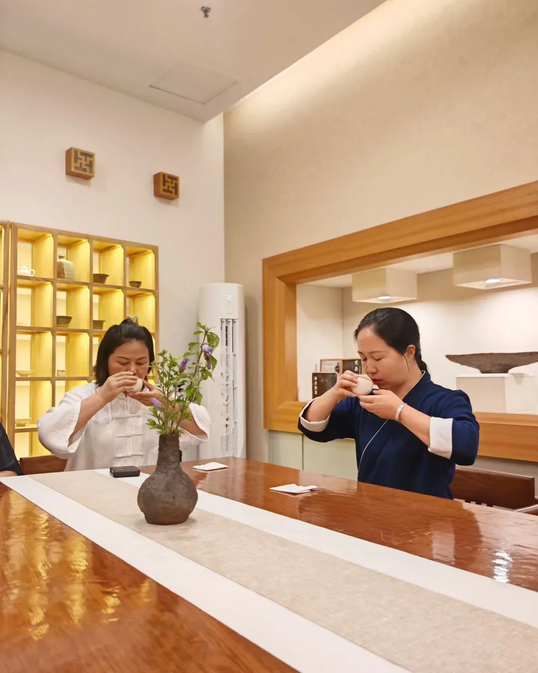 Two women across a flower vase, one in white and one in navy, both drink from small cups with their eyes closed.