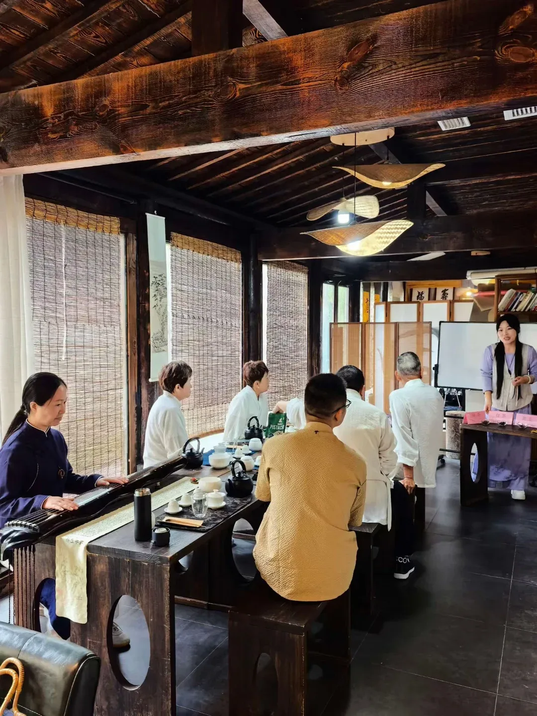In a dark timber hall with bamboo blinds, a woman plays a guqin at the end of a long tea table while students in white listen to a teacher standing by a whiteboard and small labelled tea cards.