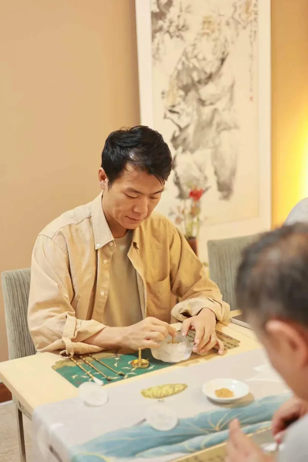 Une personne en chemise beige travaille devant un brûle-parfum en verre dépoli, sous une peinture de sage, avec des outils de laiton sur un tapis bleu-vert.