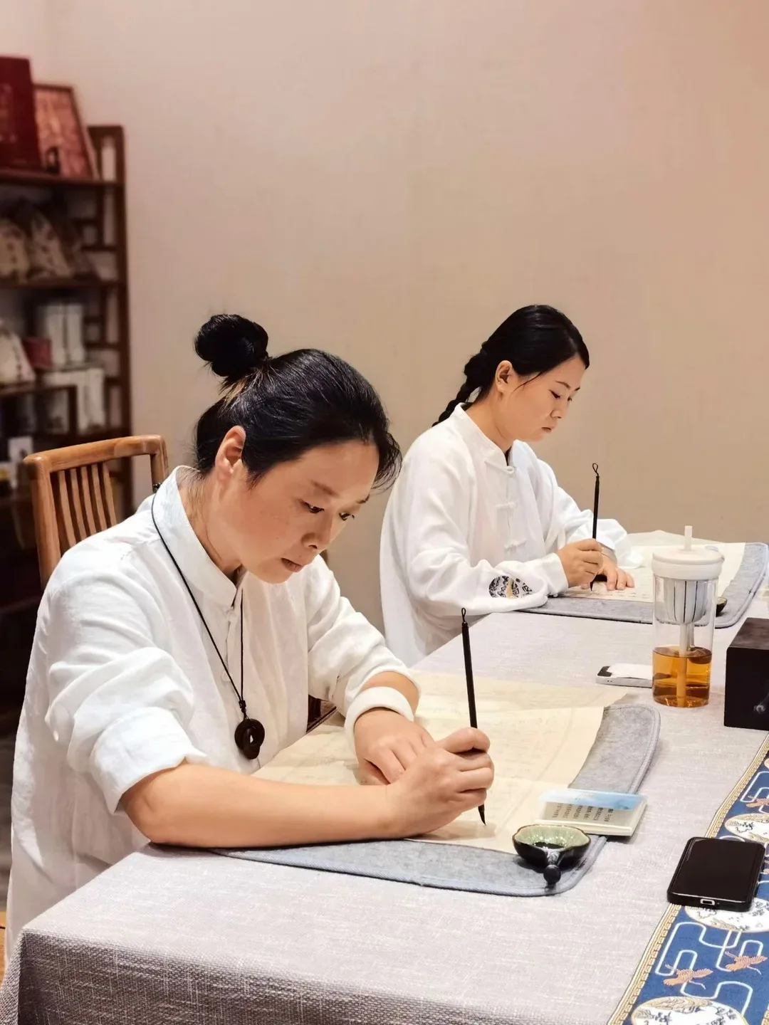 Deux femmes en blanc copient un texte au pinceau fin sur des feuilles posées sur du feutre, à une table couverte de tissu gris.