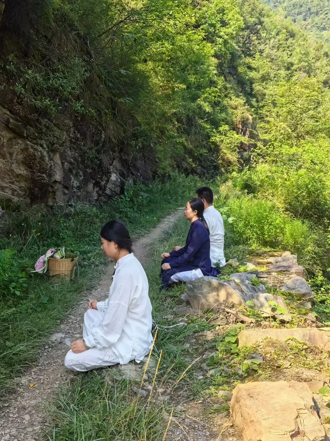 Trois personnes sont assises le long d’un sentier sous une paroi rocheuse moussue ; une personne en blanc est à genoux au premier plan, près d’un panier.