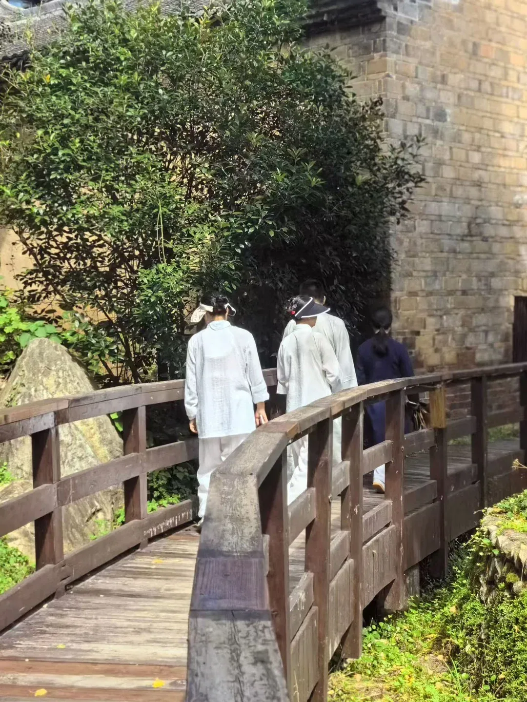Quatre personnes, trois en blanc et une en bleu marine, traversent une passerelle courbe près d’un vieux mur de briques grises et d’un arbuste dense.