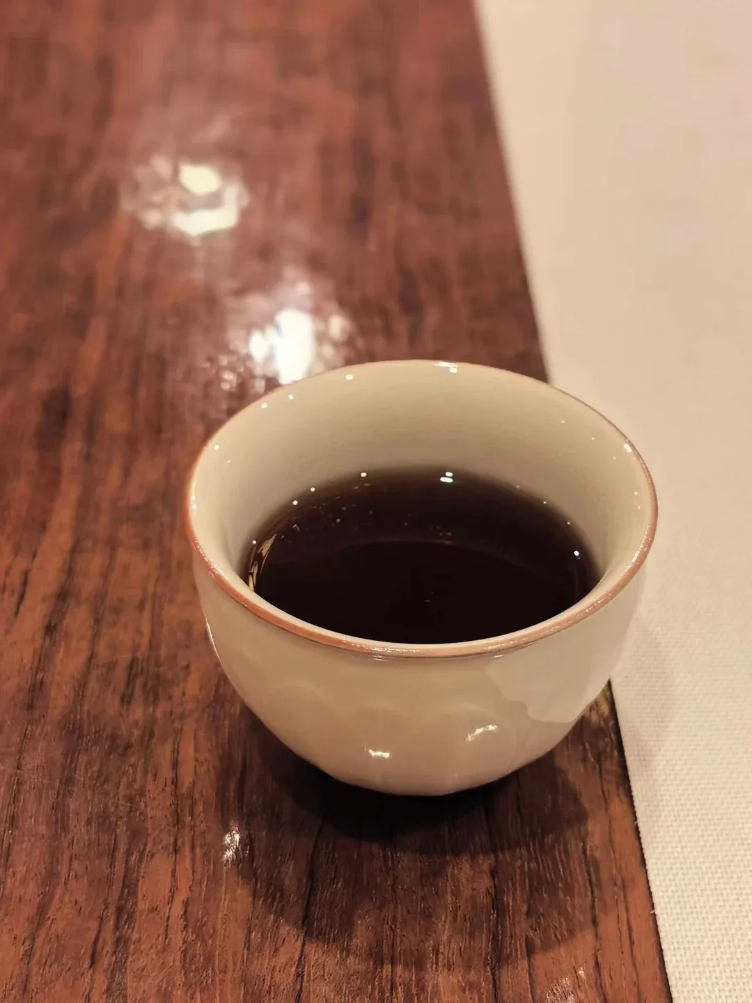 Une petite tasse crème remplie de thé rouge-brun repose sur une table de bois brillante, près d’un chemin de table en lin.