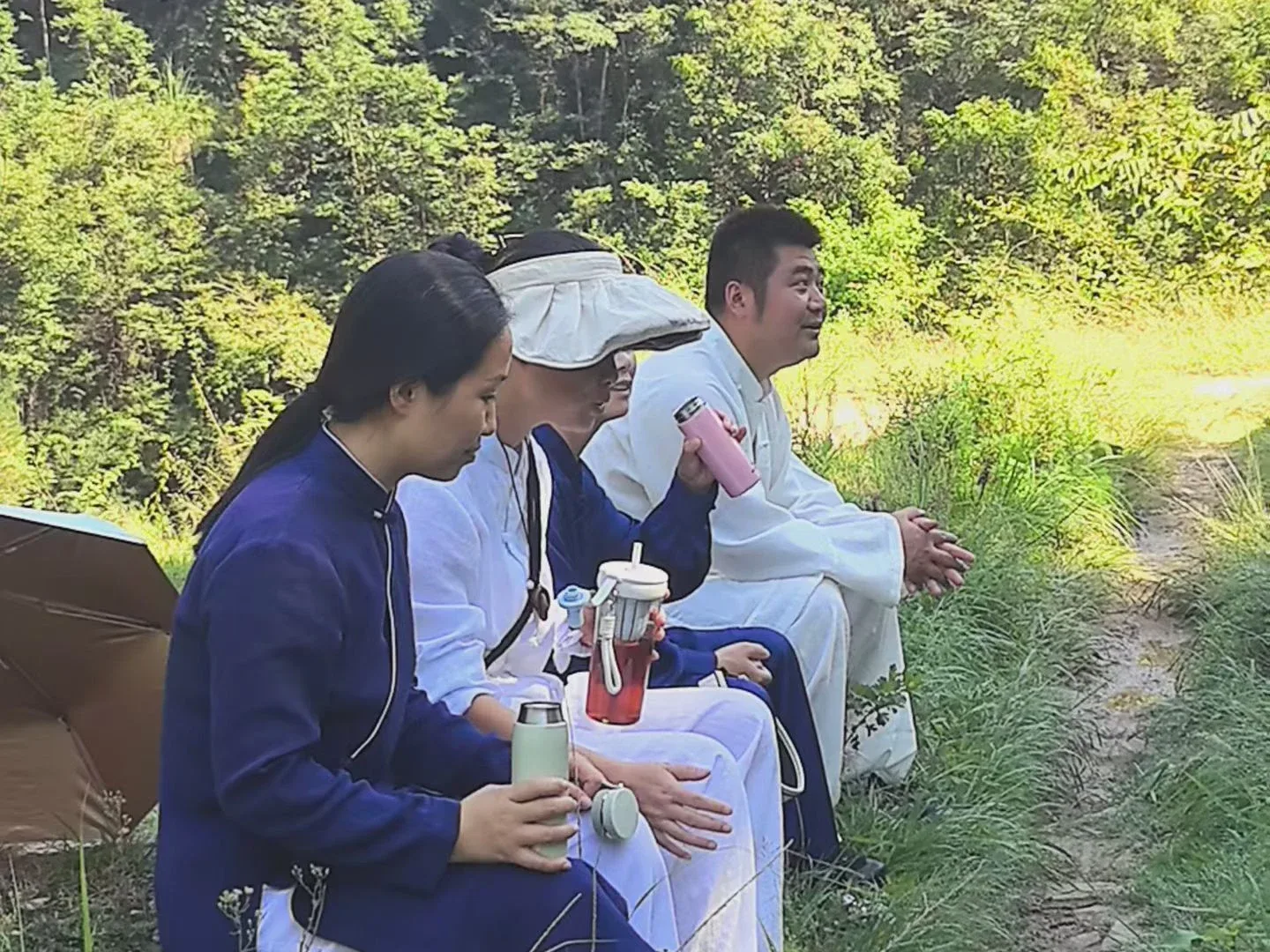 Quatre personnes en blanc et en bleu sombre s’assoient sur un talus herbeux avec leurs gourdes et un parapluie ouvert.