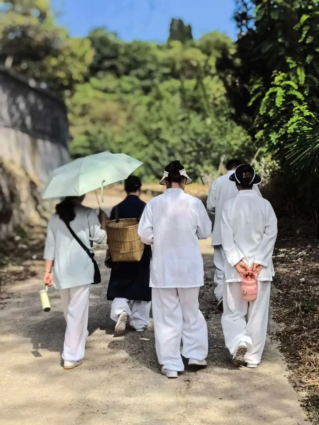 Un groupe en blanc remonte une ruelle au soleil ; une personne porte un panier de bambou, une autre tient une gourde rose derrière le dos.