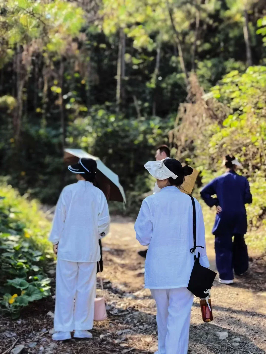 Vues de dos, des personnes en blanc et en bleu marine suivent un chemin forestier ; une bouteille pend à un sac en bandoulière.