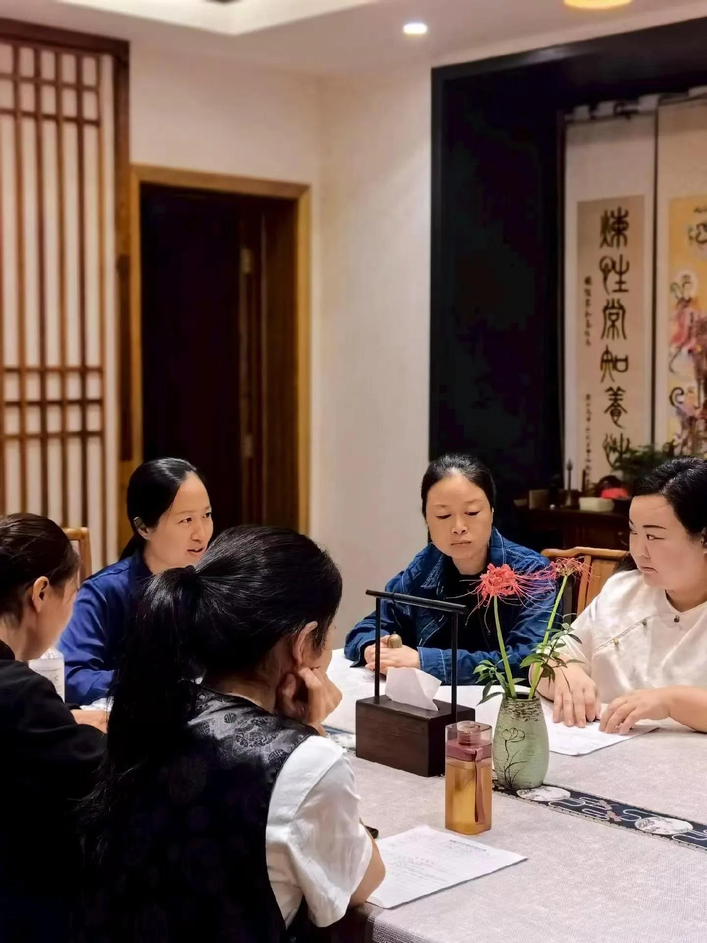 Les participants sont assis autour d’une table couverte de lin, sous un rouleau de calligraphie, avec une petite cloche et un vase de fleurs rouges.