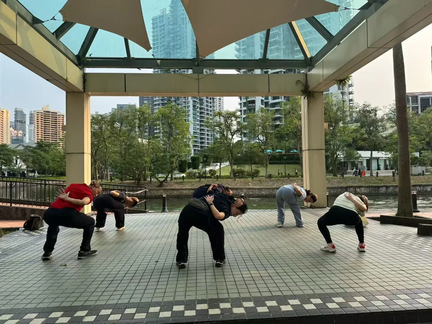 Cinq personnes se penchent et tournent le buste sous un pavillon au bord de la Singapore River, devant des immeubles résidentiels.