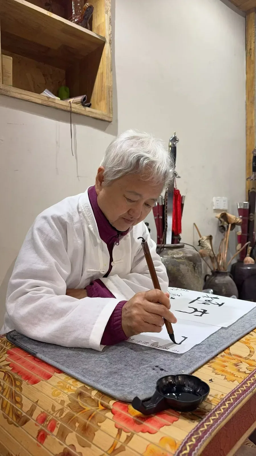 Une femme aux cheveux argentés, en veste blanche sur des vêtements prune, trace des caractères avec un pinceau vertical sur une table couverte d’une natte.