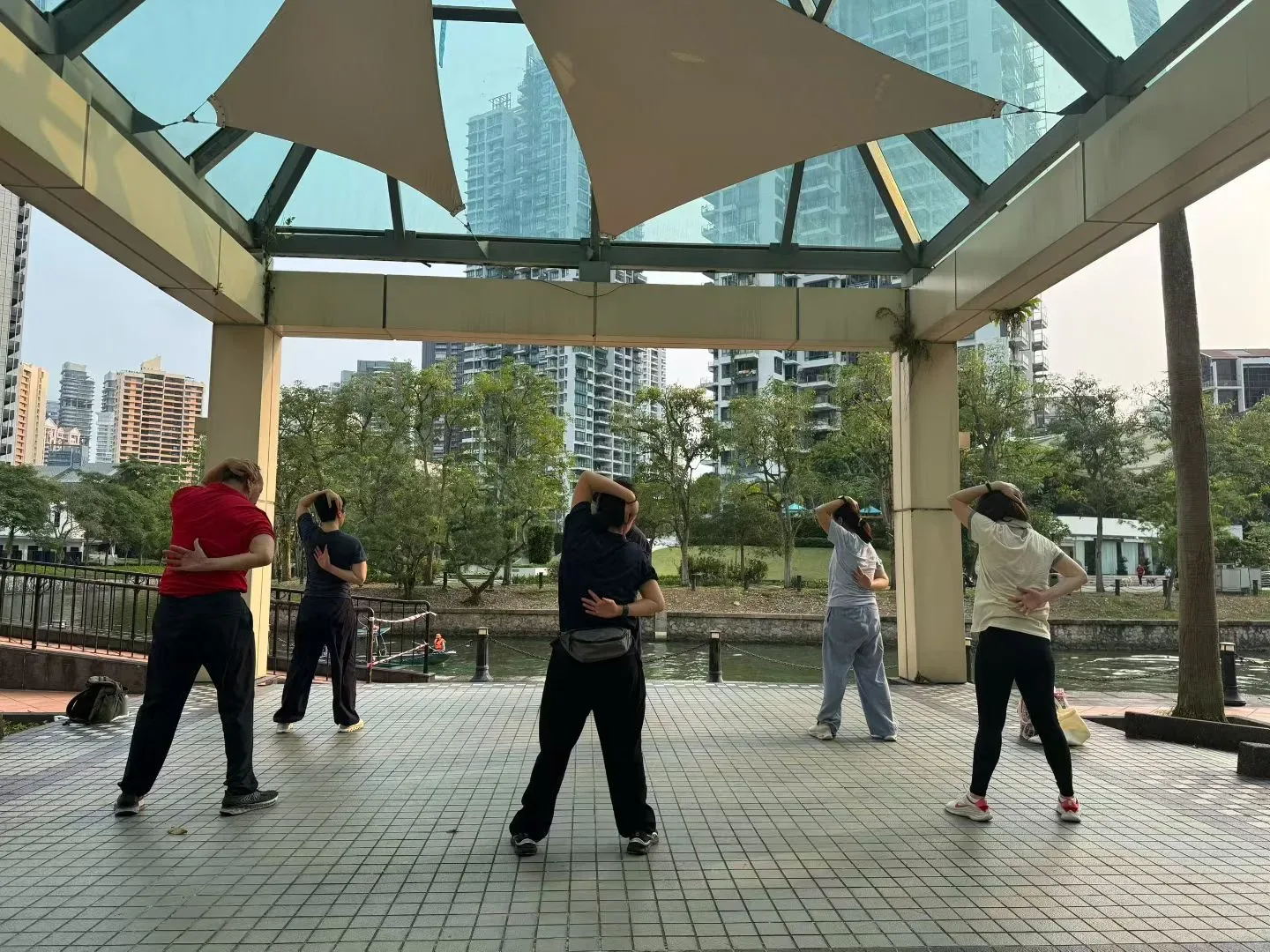 Cinq personnes tournent le buste sous un pavillon vitré face à la Singapore River, une main derrière la tête et l’autre au bas du dos.