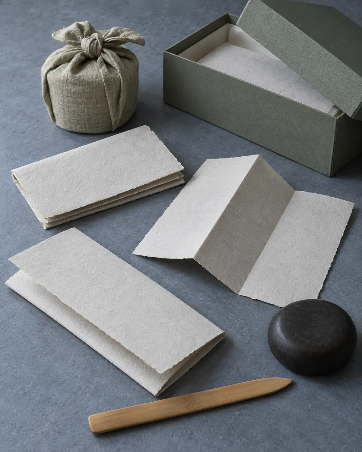 Papiers pliés vierges, boîte à thé enveloppée de tissu, boîte d’archives, couteau à papier en bambou et presse-papiers sombre sur une table gris froid