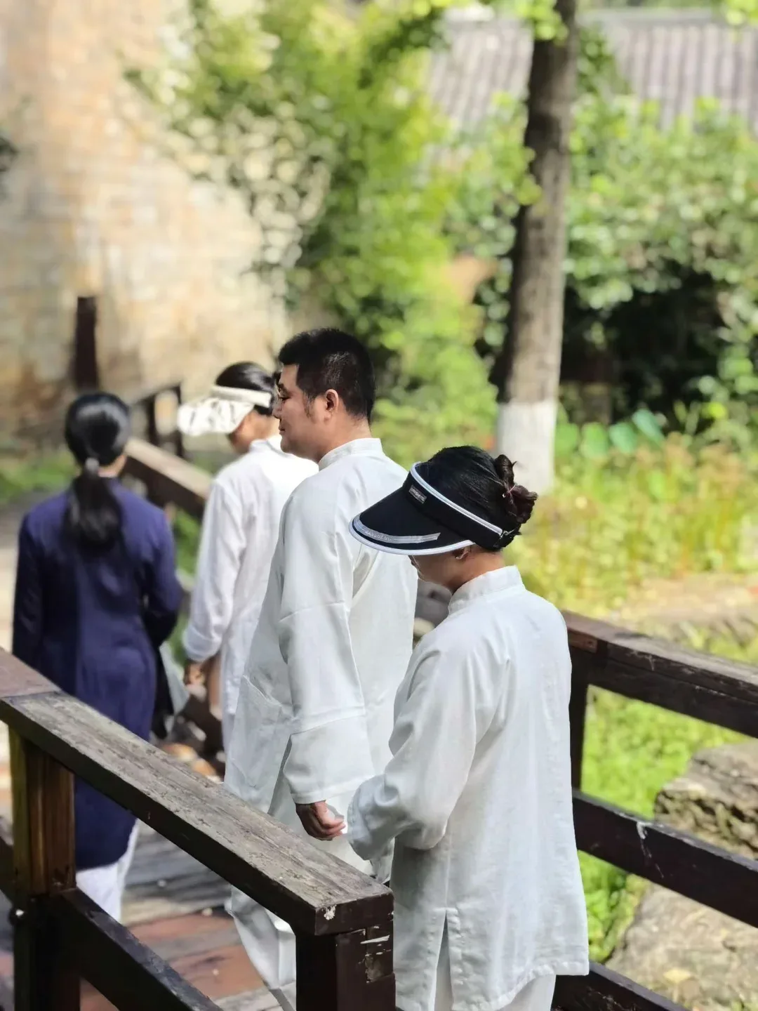 Quatre personnes en blanc et en robes sombres traversent en file une passerelle de bois bordée de verdure.