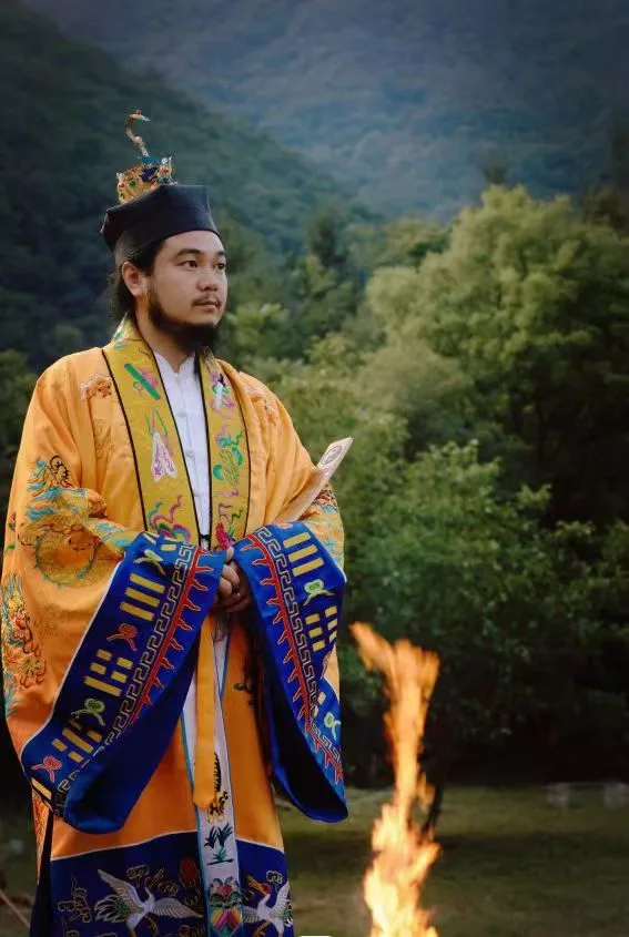 Teacher Facheng in ceremonial Daoist ritual vestments, holding a document, beside a fire in a forest clearing.