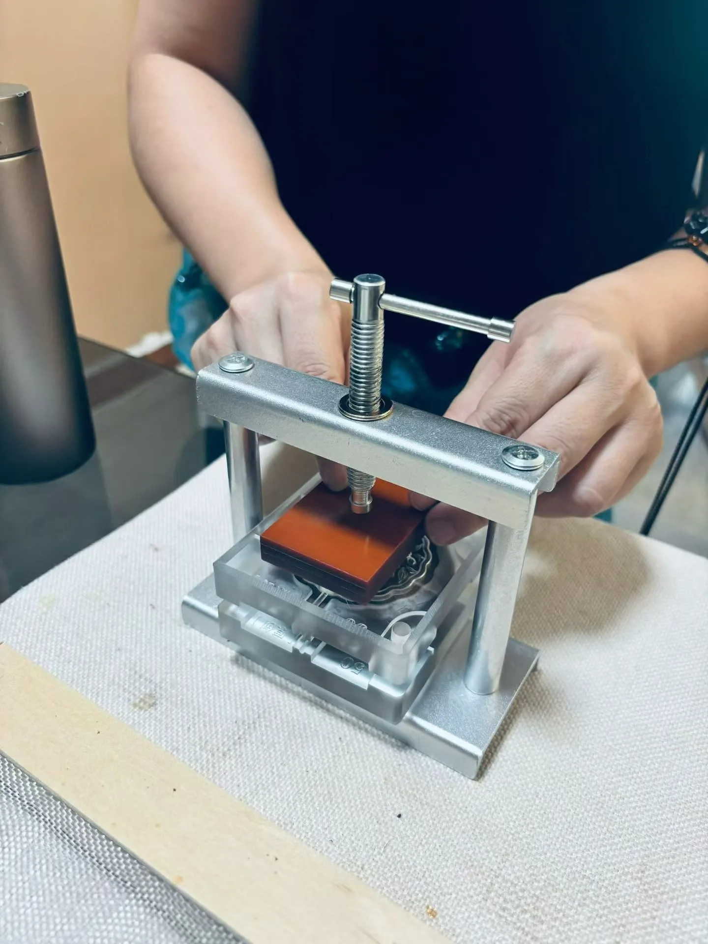 Des mains tournent la poignée d’une petite presse à vis métallique contenant un moule et une cale de bois rouge.