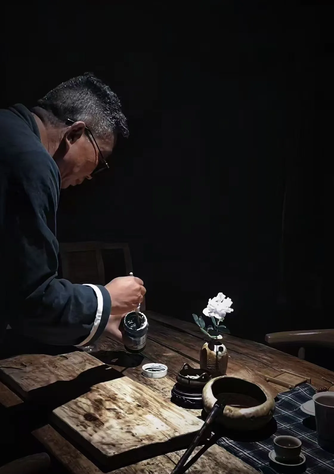 Chen Haichao leans over a wooden studio table by lamplight, brush in hand beside an ink jar and a white peony.
