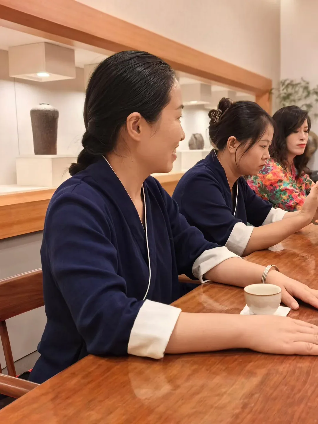 Three women seated along a wooden table talk and smile after tea, one gesturing as she speaks, a small cup on its paper coaster in front of her.