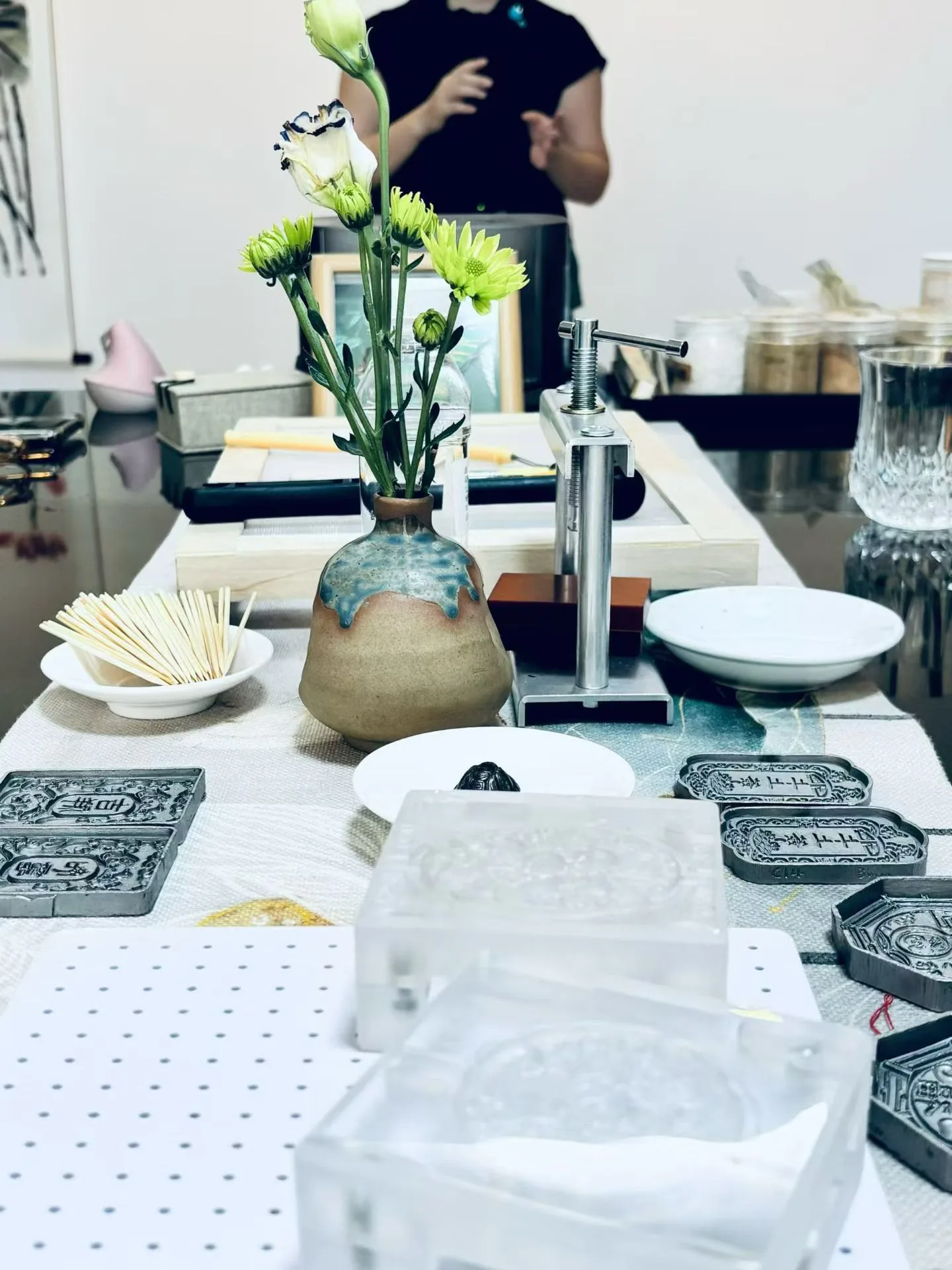 La table de l’atelier réunit un vase de chrysanthèmes verts, une presse à vis, des moules transparents et des formes métalliques gravées.
