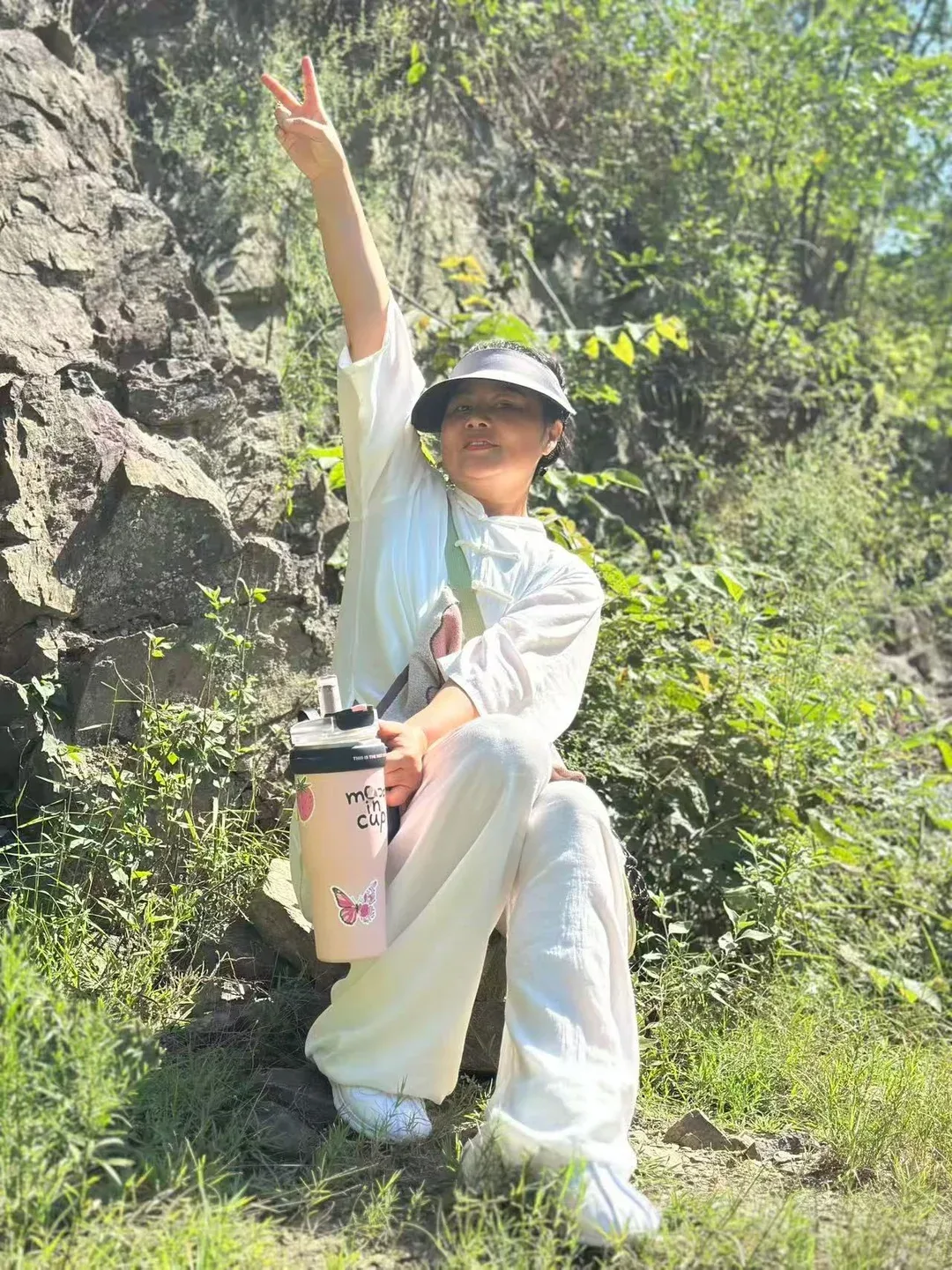 Une femme en blanc s’appuie contre un rocher et lève deux doigts vers l’appareil ; une grande bouteille d’eau repose près de son genou.
