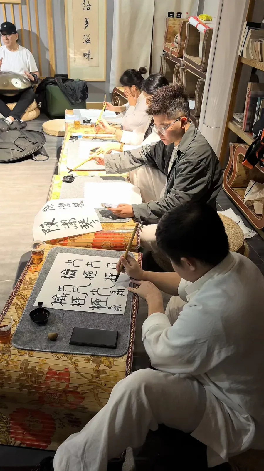 Des élèves pratiquent la calligraphie sur des tapis de feutre à des tables basses en bambou ; une personne tient un handpan à l’arrière-plan.