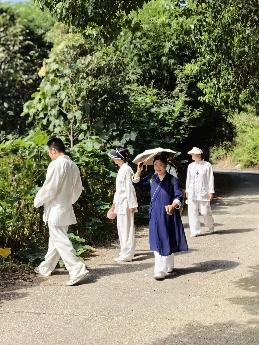 Cinq personnes en blanc et en bleu marchent sur une route ombragée ; l’une tient un parapluie clair et d’autres portent des visières.