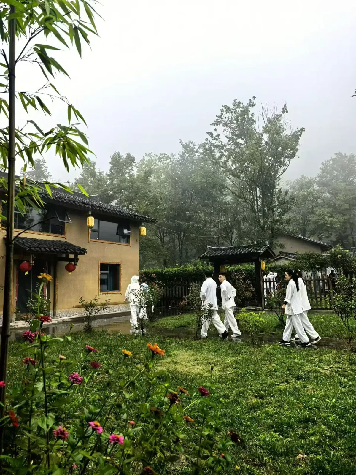 Des personnes en tenue blanche traversent une pelouse humide dans la brume, près d’une maison de terre ornée de lanternes, derrière des bambous et des zinnias.