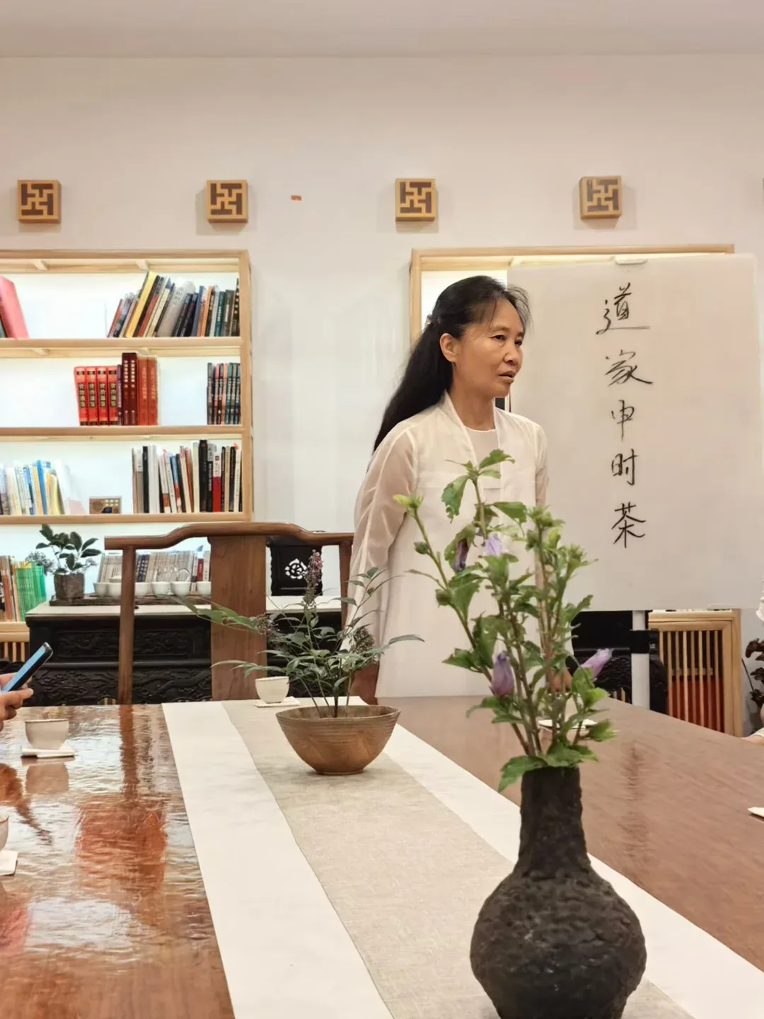 A woman with long dark hair in a white gown speaks beside a sheet brushed with the characters 道家申时茶; a vase of purple hibiscus stands on the table before her.