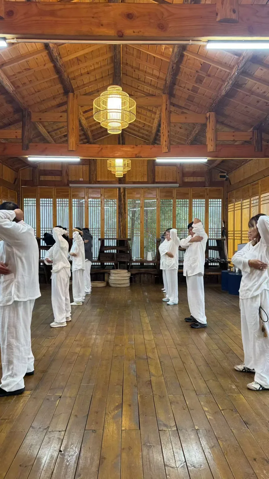 Dans une salle en bois, deux rangées de personnes en blanc mobilisent les bras et la nuque sous des lanternes de bambou.